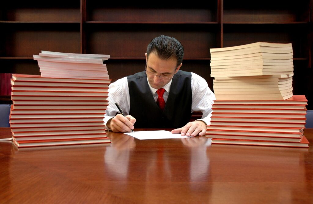 A man in a suit writes on paper at a table surrounded by stacks of books. The image highlights dedication to scholarly work or research.
