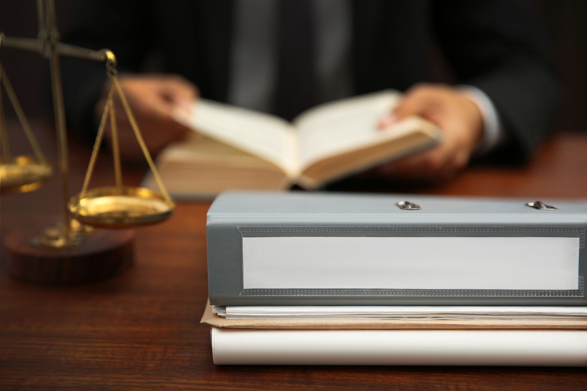 Legal documents and a scale of justice on a desk, with a blurred figure reading a book, emphasize legal practice and research.