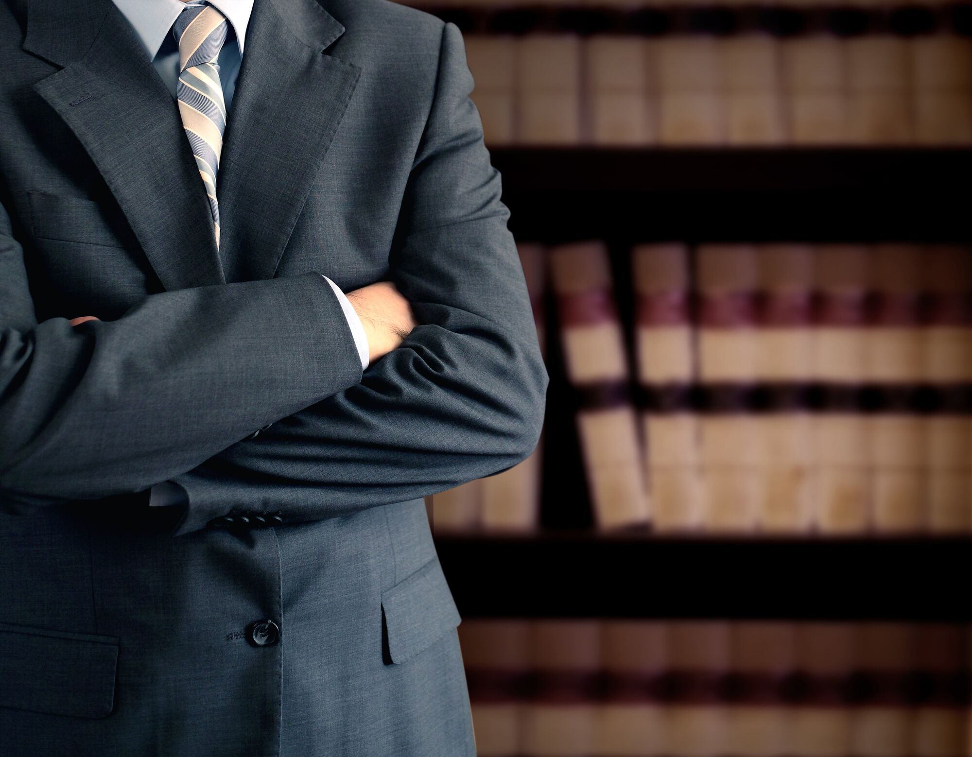 A confident person in a suit stands with crossed arms in front of a blurred bookshelf filled with law books, suggesting a legal context.